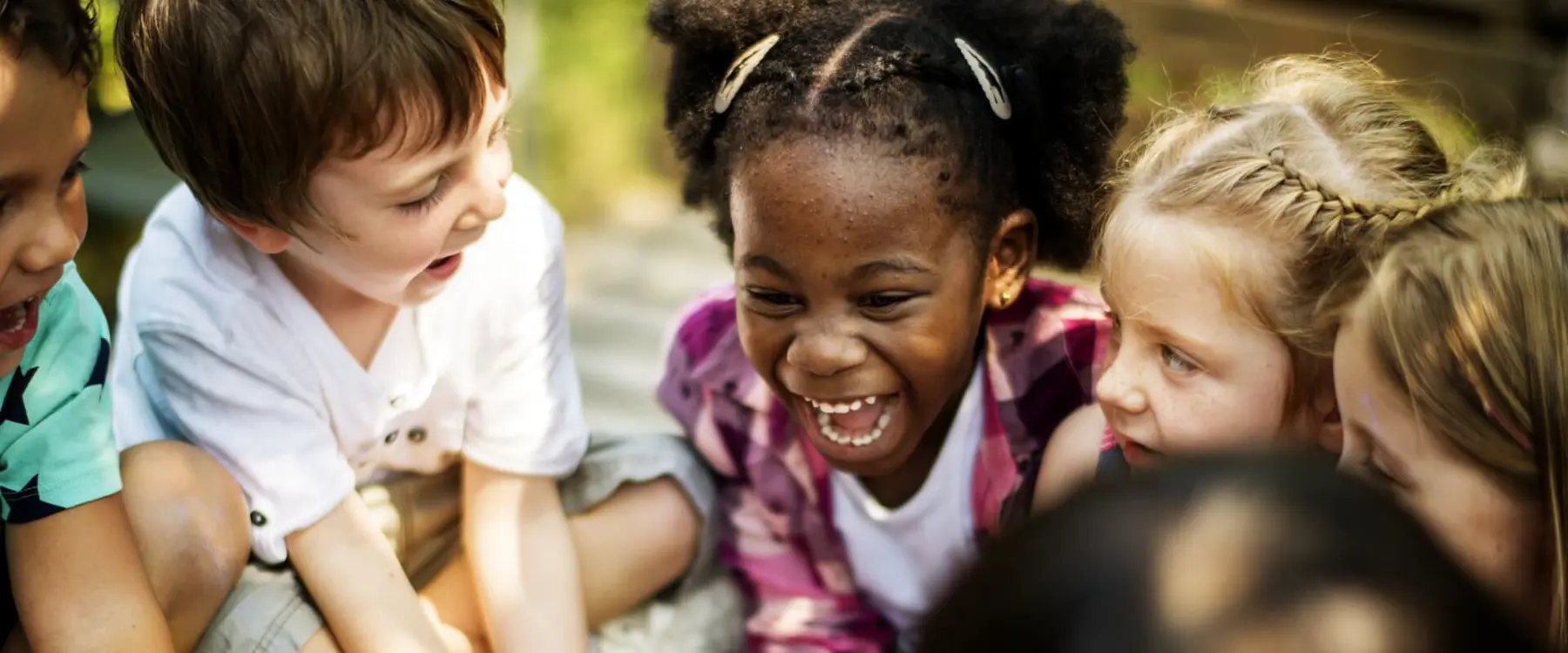 A group of young children sit close together outdoors, smiling and laughing as they look downward at something out of frame.