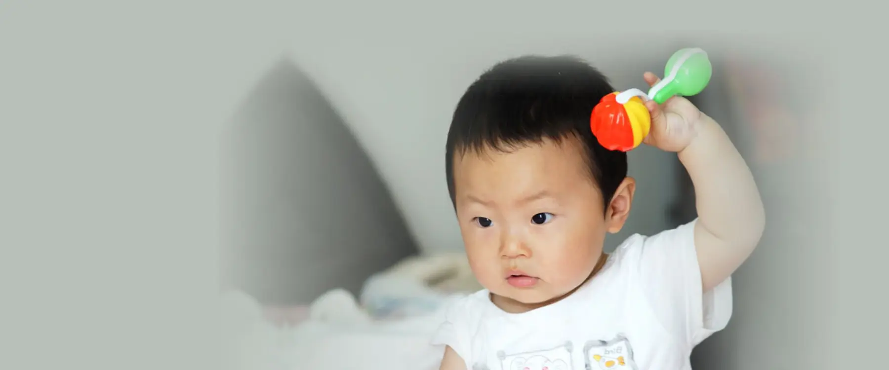 A baby in a white shirt holds a colorful rattle up near their head while looking downward.