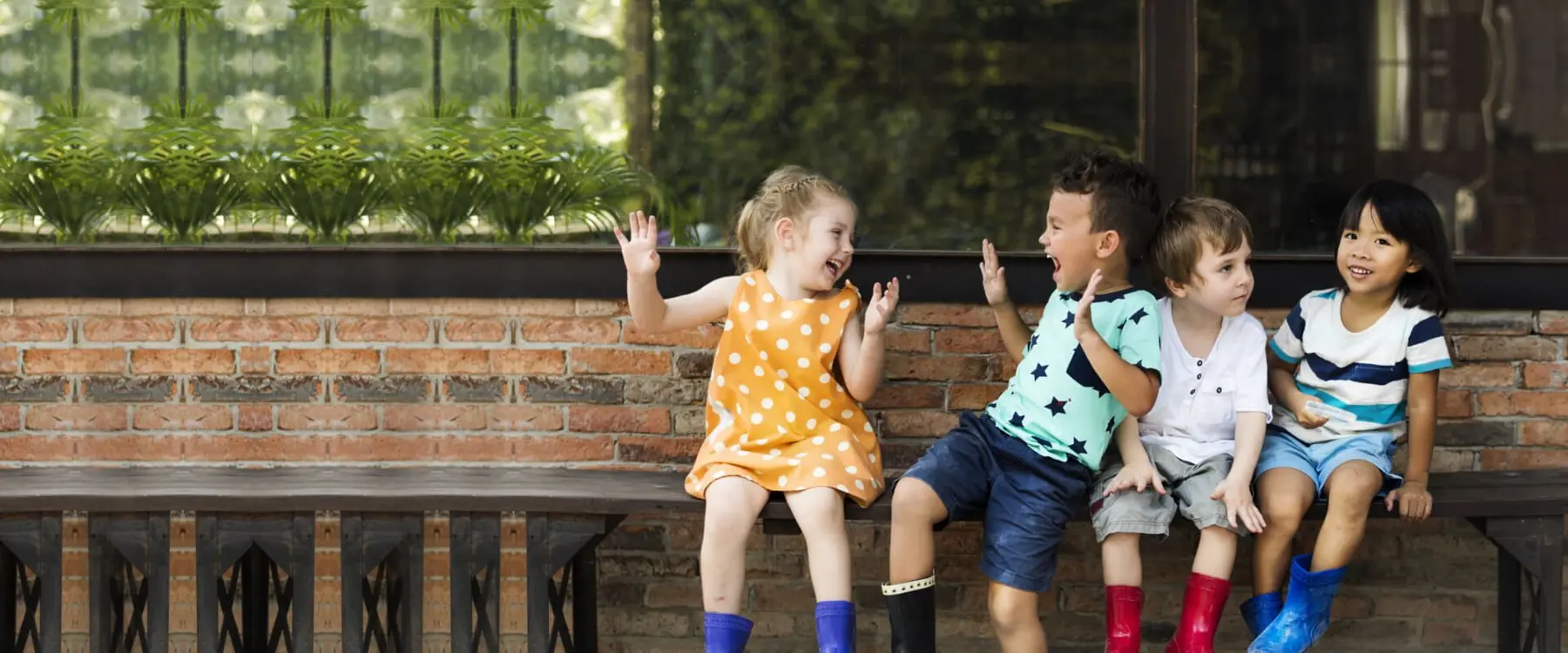 Four children sit on a wooden bench against a brick wall, laughing and gesturing, with three wearing rain boots.