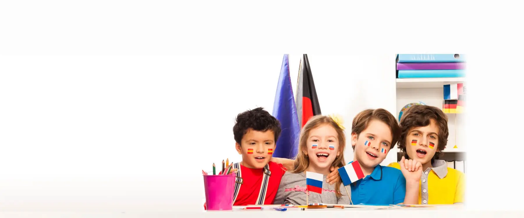 Four children sit at a table with painted flags on their cheeks, holding small flags and smiling, with a pink cup of pencils and a shelf of binders behind them.