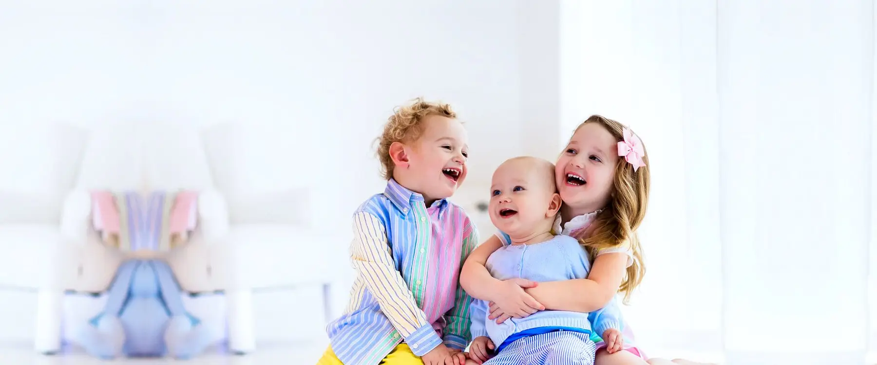 Two older children sit and hug a baby on a light floor, all smiling, with a blurred white chair and hanging fabric in the background.