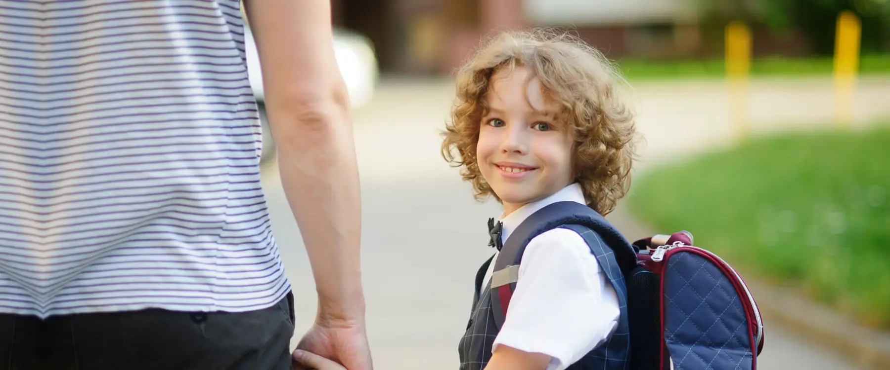 A child with curly hair wearing a white shirt, tie and backpack smiles while holding the hand of an adult in a striped shirt.