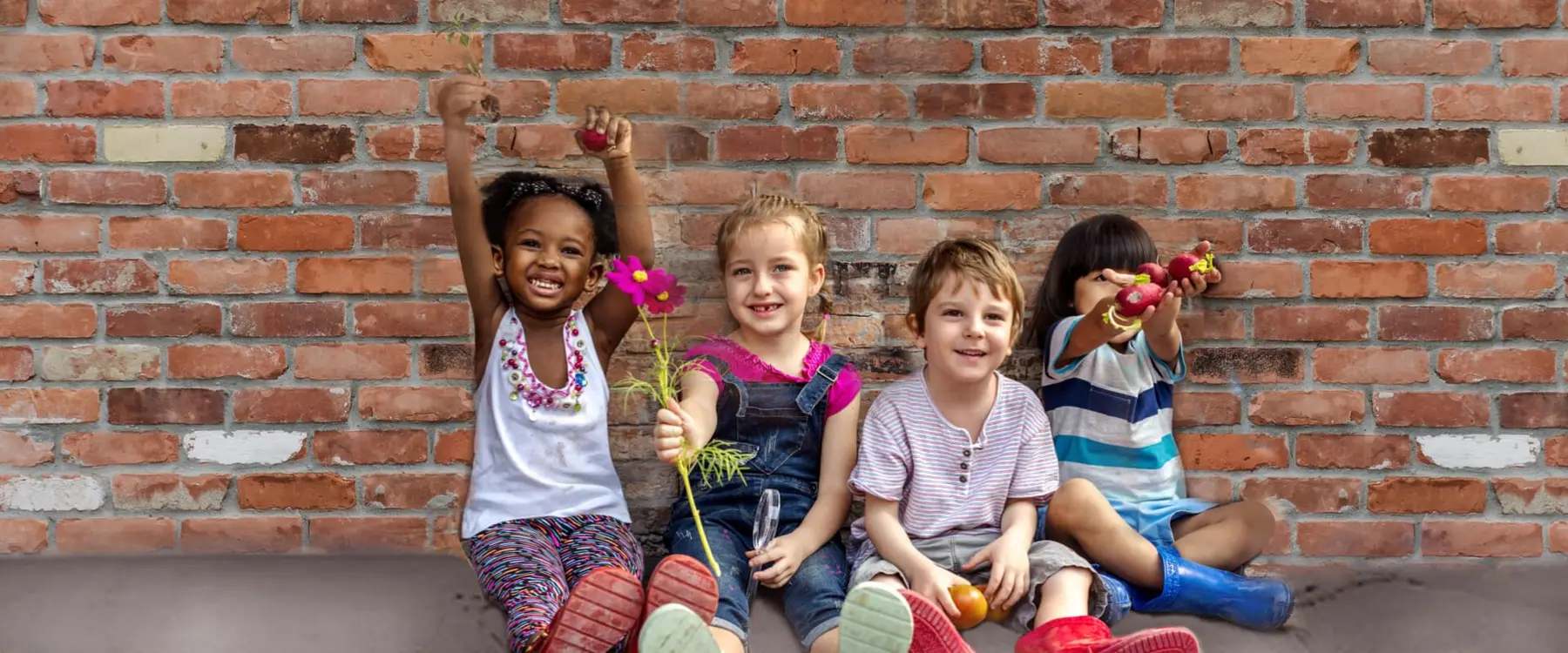 Four children sit against a brick wall, one girl raising a flower and another holding a flower, while two boys hold small fruits.