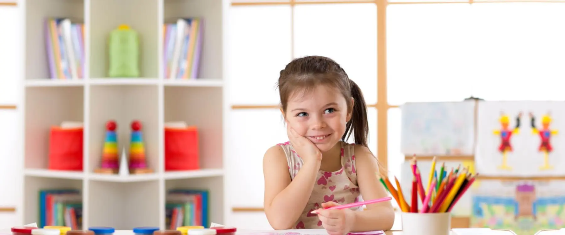 A young girl rests her chin on her hand while holding a pink pencil over paper at a table with a cup of colored pencils and a shelf of books behind her.