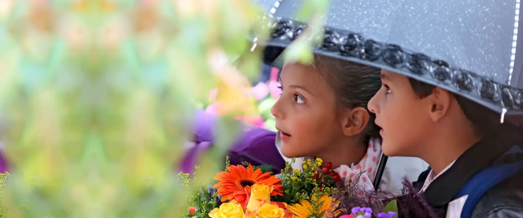A boy and a girl stand close together under a black umbrella, looking toward the left past a bouquet of orange and yellow flowers.