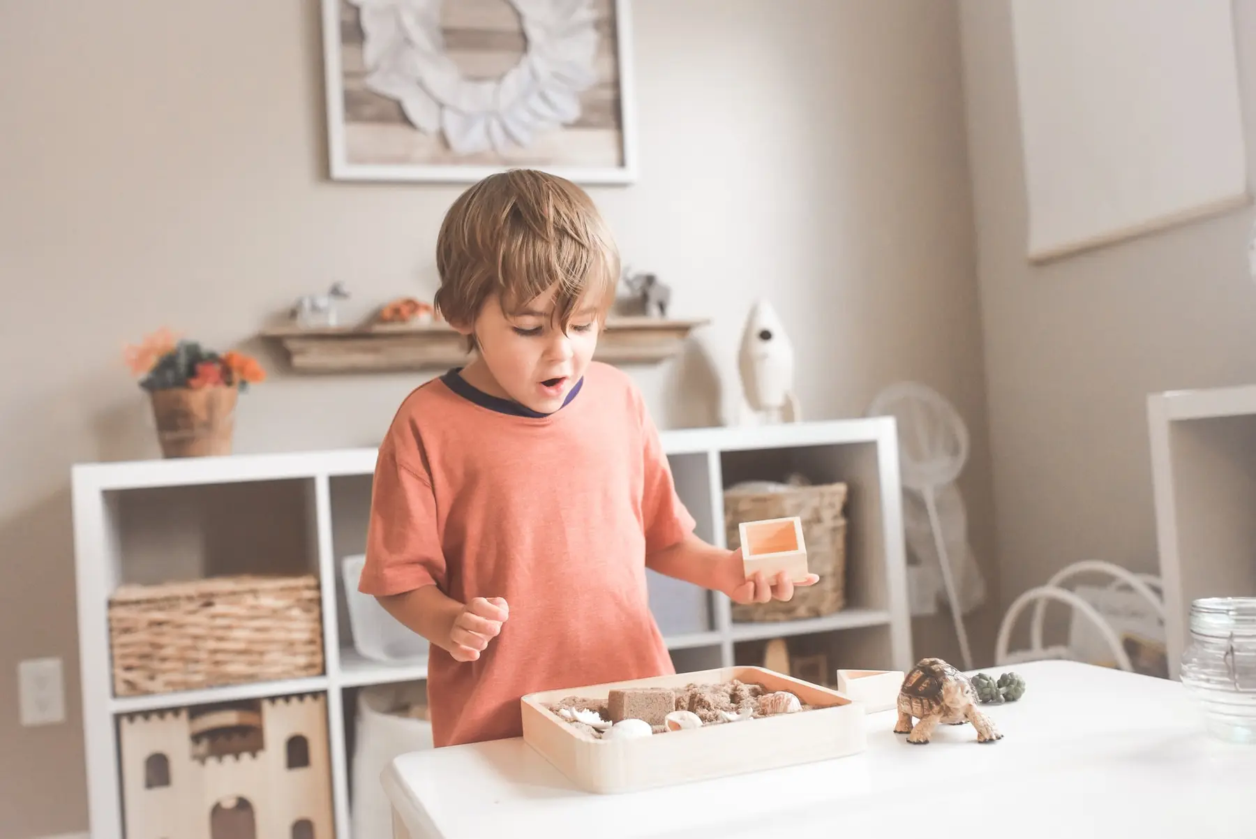 A young boy in an orange shirt stands at a white table holding a small wooden box above a tray of sand and toy dinosaurs.