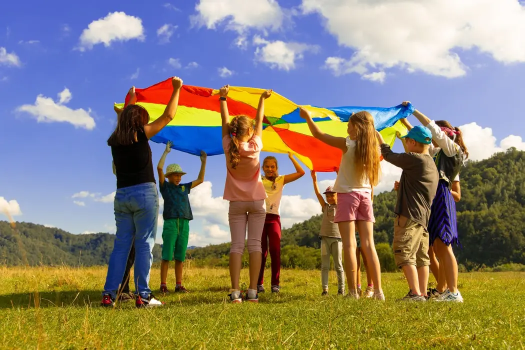 A group of children and one adult stand in a grassy field holding the edges of a large multicolored parachute up against a blue sky with clouds.