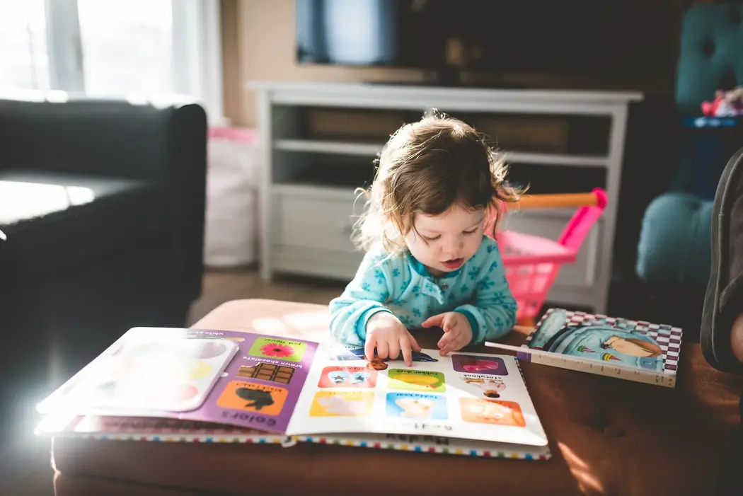 A young child in a blue patterned top sits on a brown couch and points at an open picture book resting on the cushion.