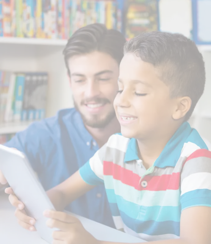 A man and a boy sit together looking at a tablet screen in front of shelves of books.