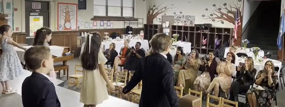 Children in formal clothing stand in a line facing seated adults who are taking photos with phones and cameras in a classroom with wooden cubbies and tree murals.