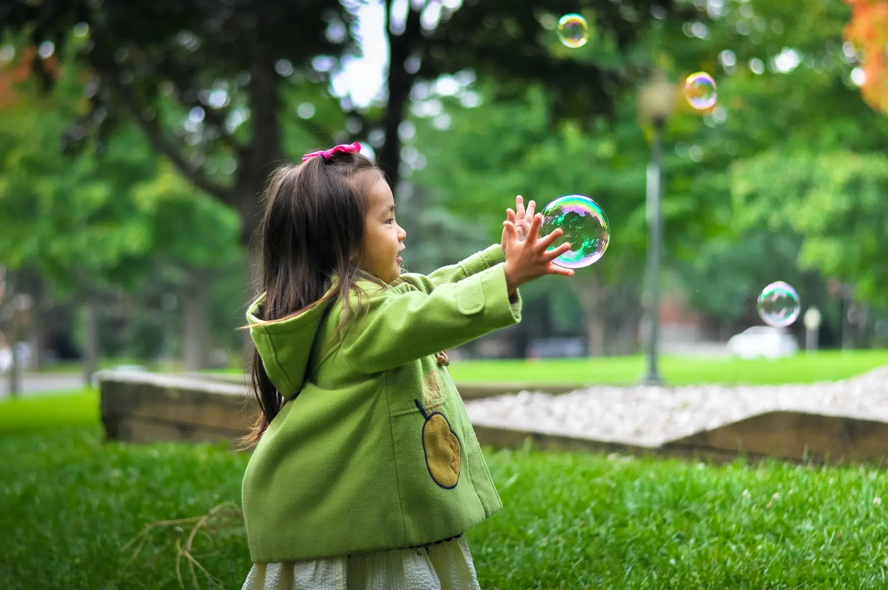A young girl in a green coat reaches toward a large soap bubble floating in a park.
