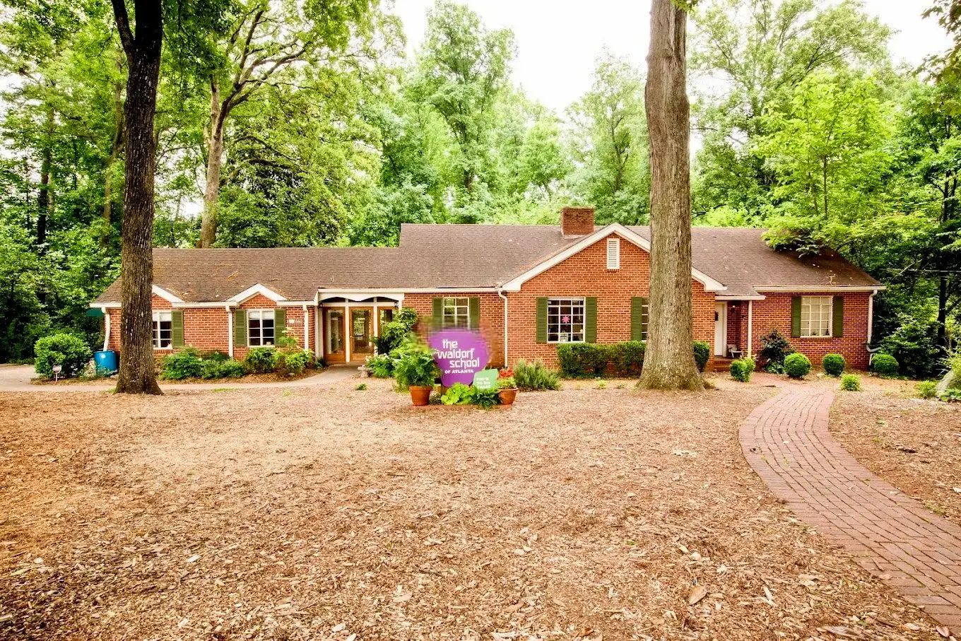 A single-story red brick building with a brown shingled roof and green shutters sits behind a large mulched yard with a purple sign reading 'The Goddard School' in front.