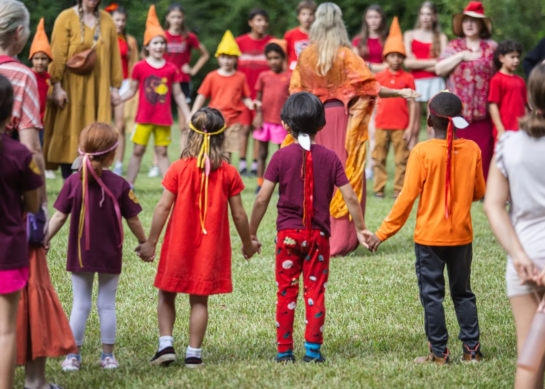Children and adults stand in a circle on grass holding hands, with several children wearing paper cone hats and one adult in a long yellow dress.