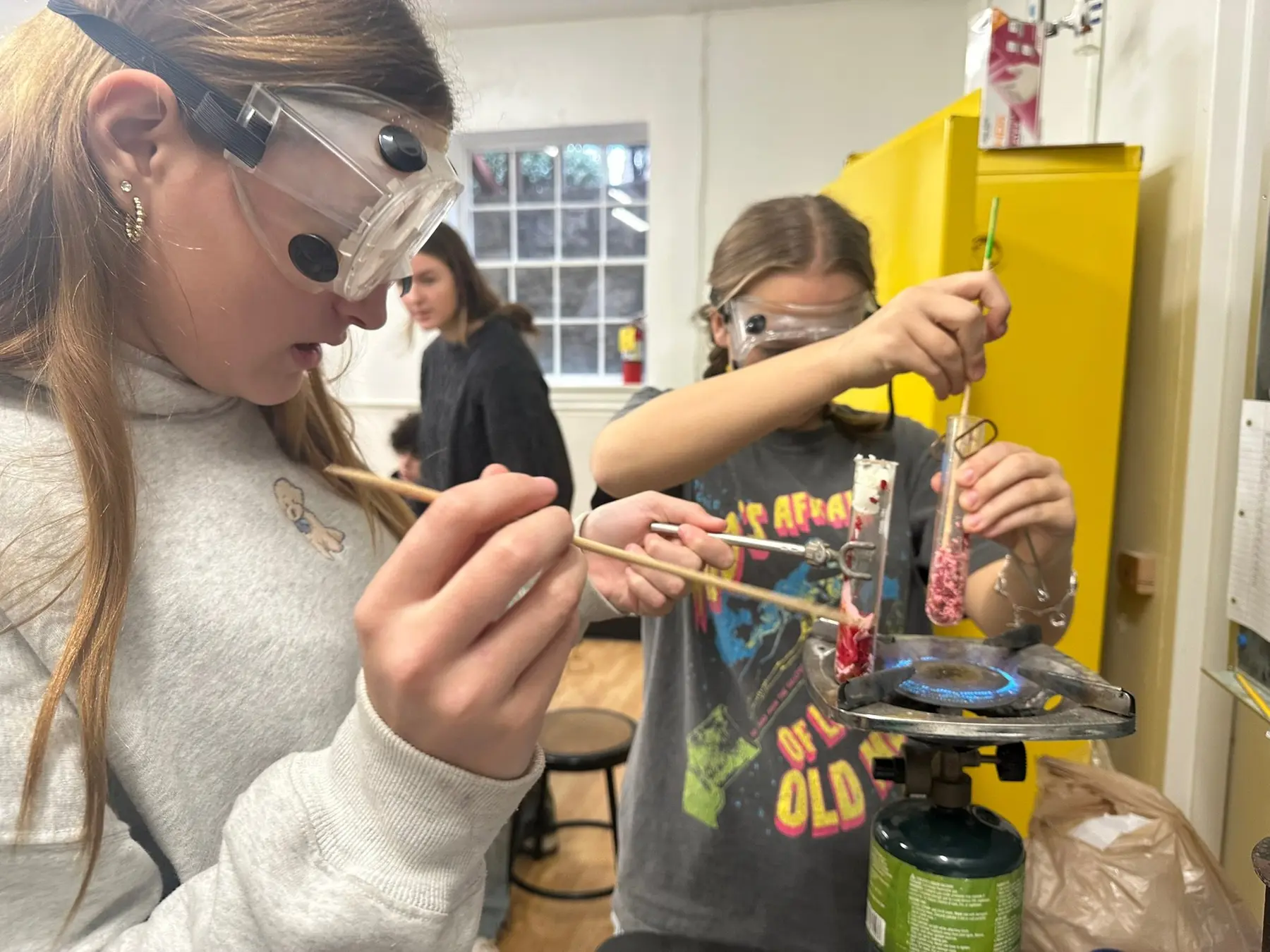Two girls wearing safety goggles hold wooden sticks and a test tube over a lit Bunsen burner on a lab table while a third person stands in the background.