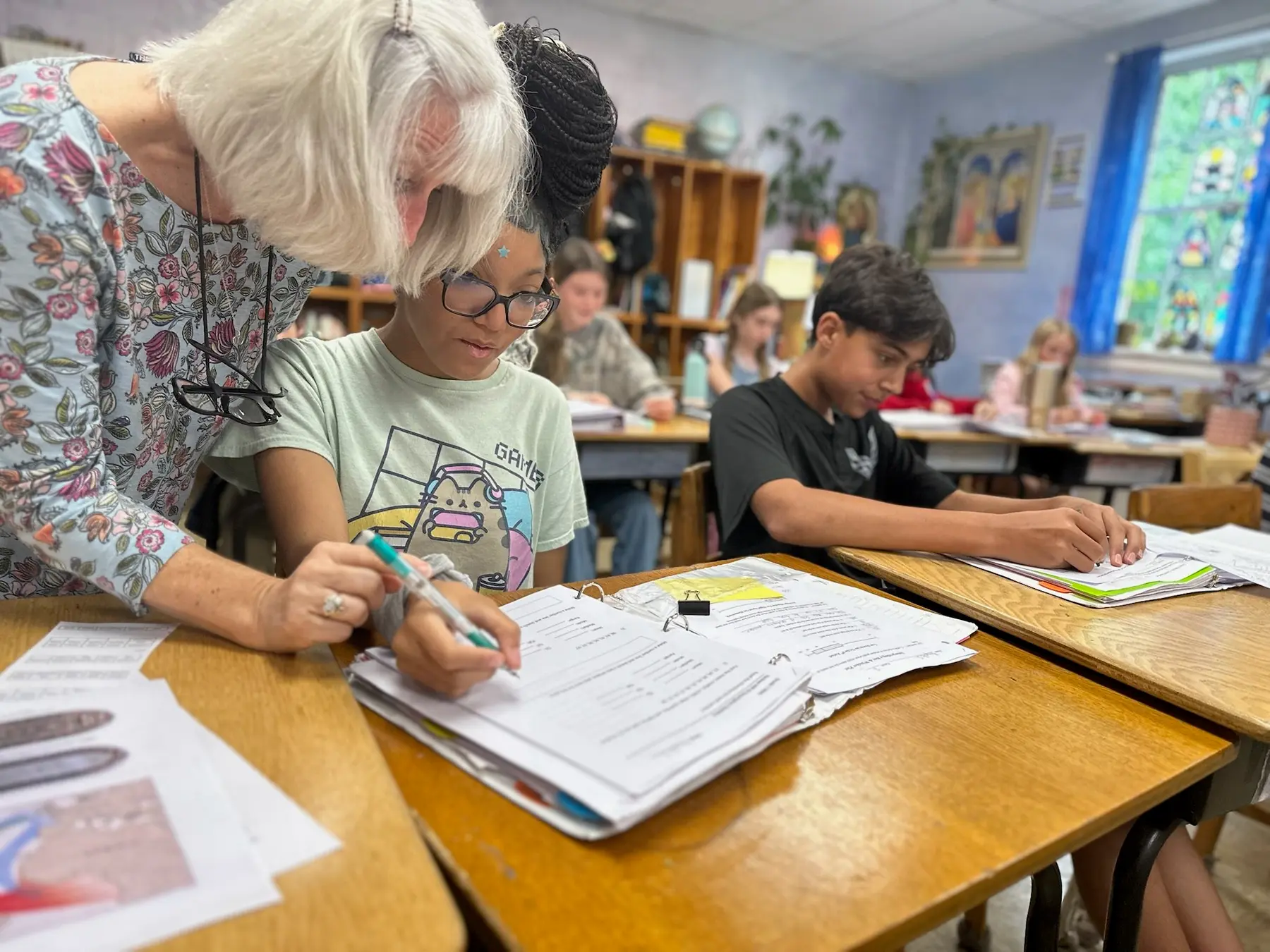 An adult with short white hair leans over a desk pointing at a binder with a pen while a student in a light green shirt watches, and other students work at desks nearby.