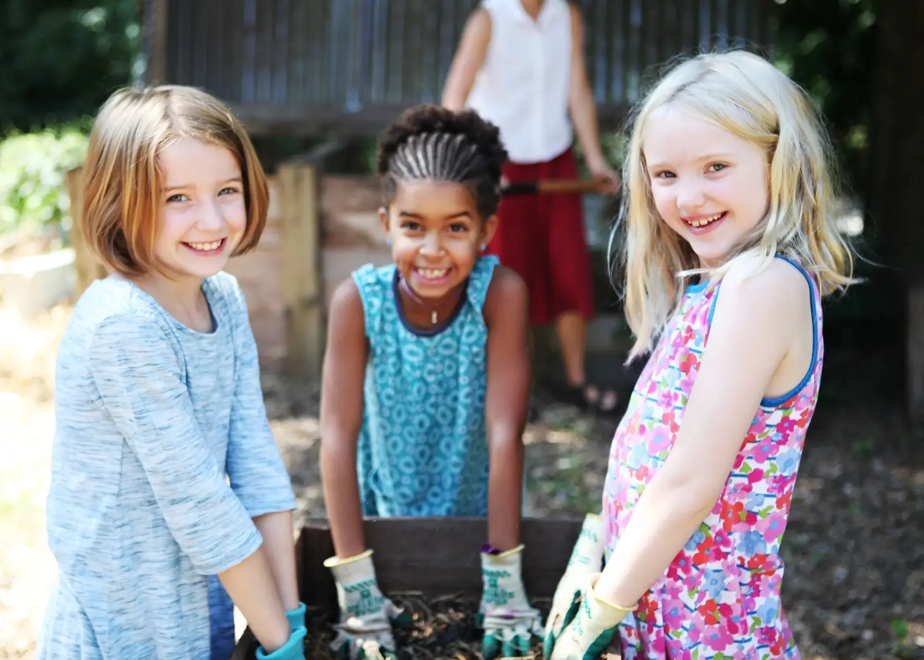 Three girls stand around a wooden planter box wearing gardening gloves while an adult in the background holds a tool.