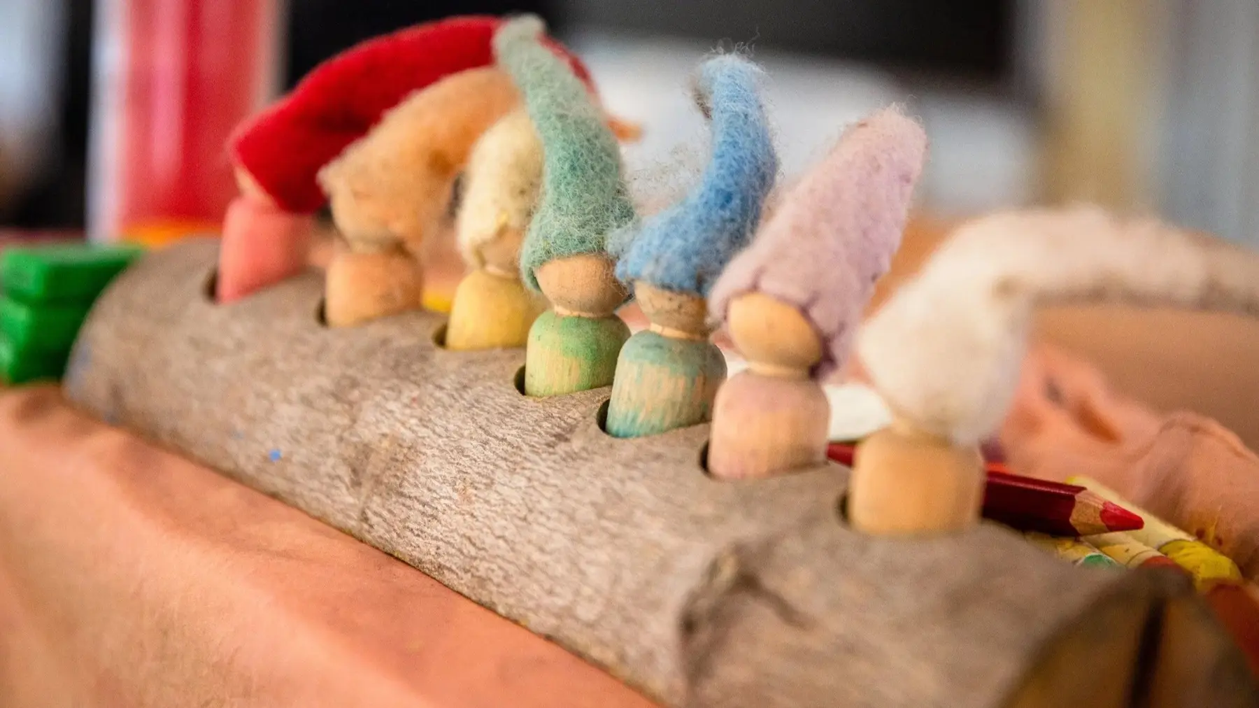 A row of small wooden peg dolls with colorful felt hats sits inside holes drilled into a cardboard tube resting on a table.