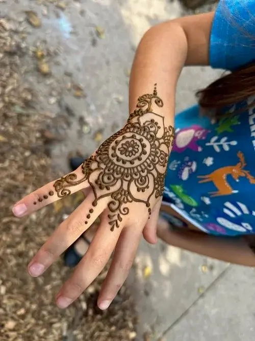 A child's hand with brown henna designs rests on a paved surface, wearing a blue shirt with animal graphics.