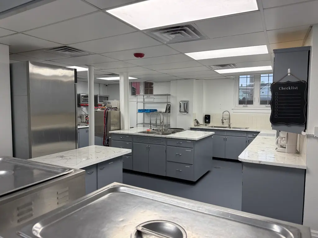 A commercial kitchen with stainless steel appliances, gray cabinets, marble-patterned countertops, and a hanging checklist sign.