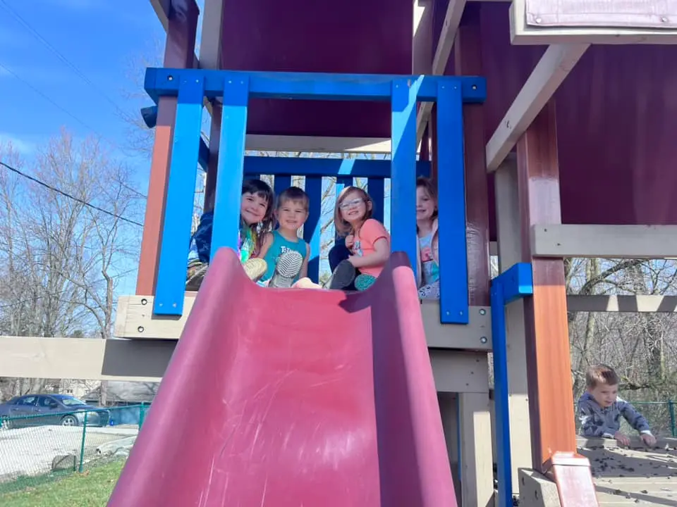 Four children sit at the top of a playground slide while a fifth child climbs on the structure to the right.