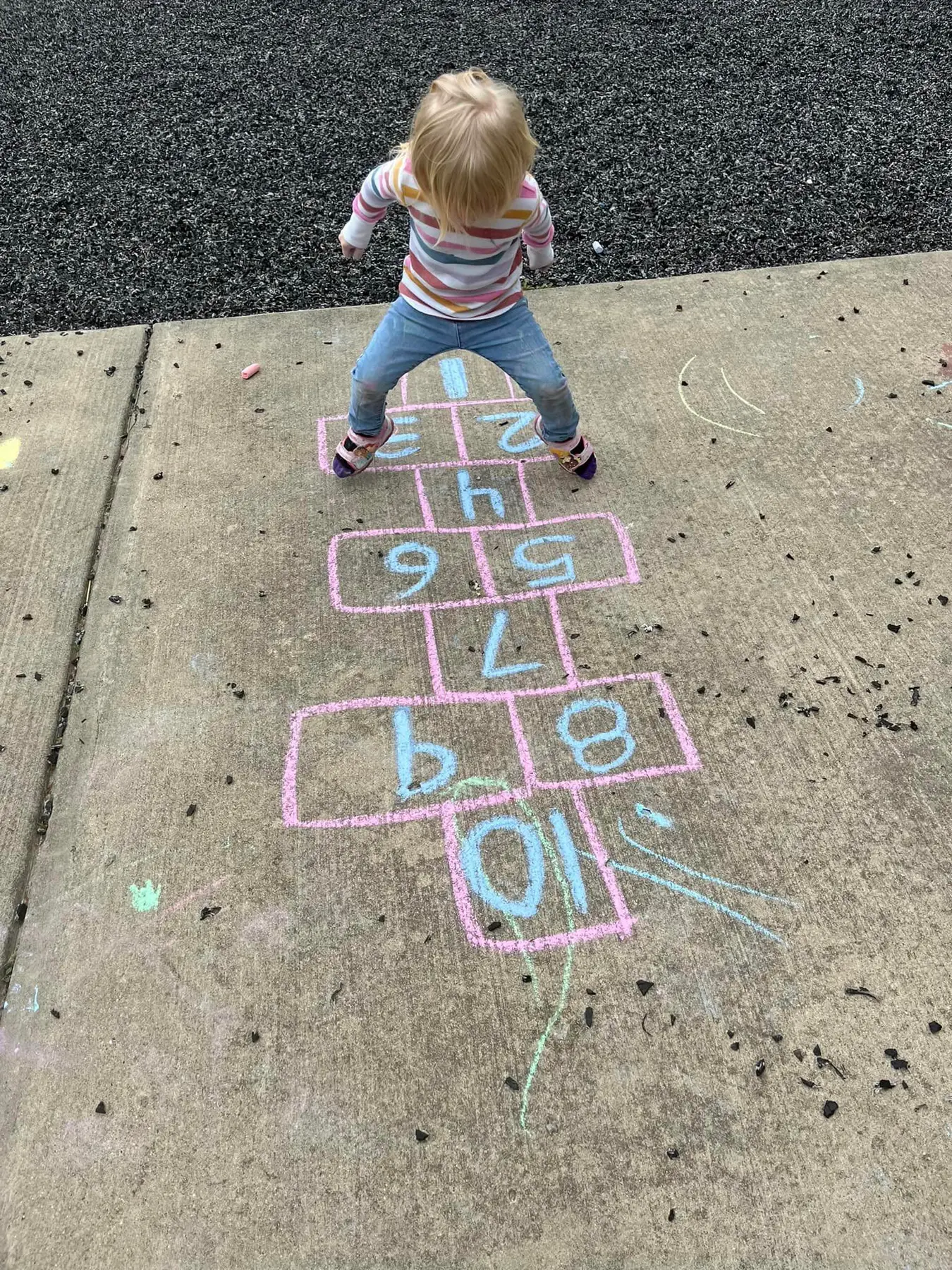 A young child with blonde hair stands on a chalk-drawn hopscotch grid on a concrete surface, facing away from the camera.