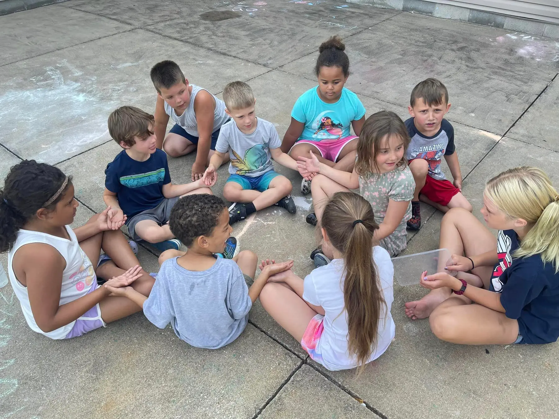 Ten children sit in a circle on a concrete surface, facing inward and gesturing with their hands.