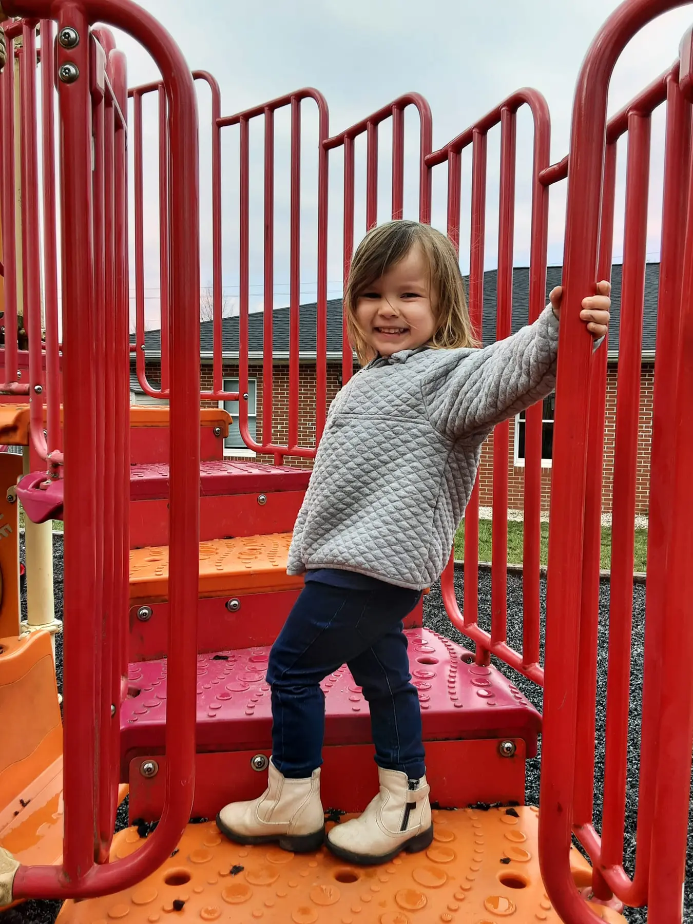 A smiling child in a gray quilted jacket, blue jeans, and white boots stands on red and orange playground steps, holding a red railing.