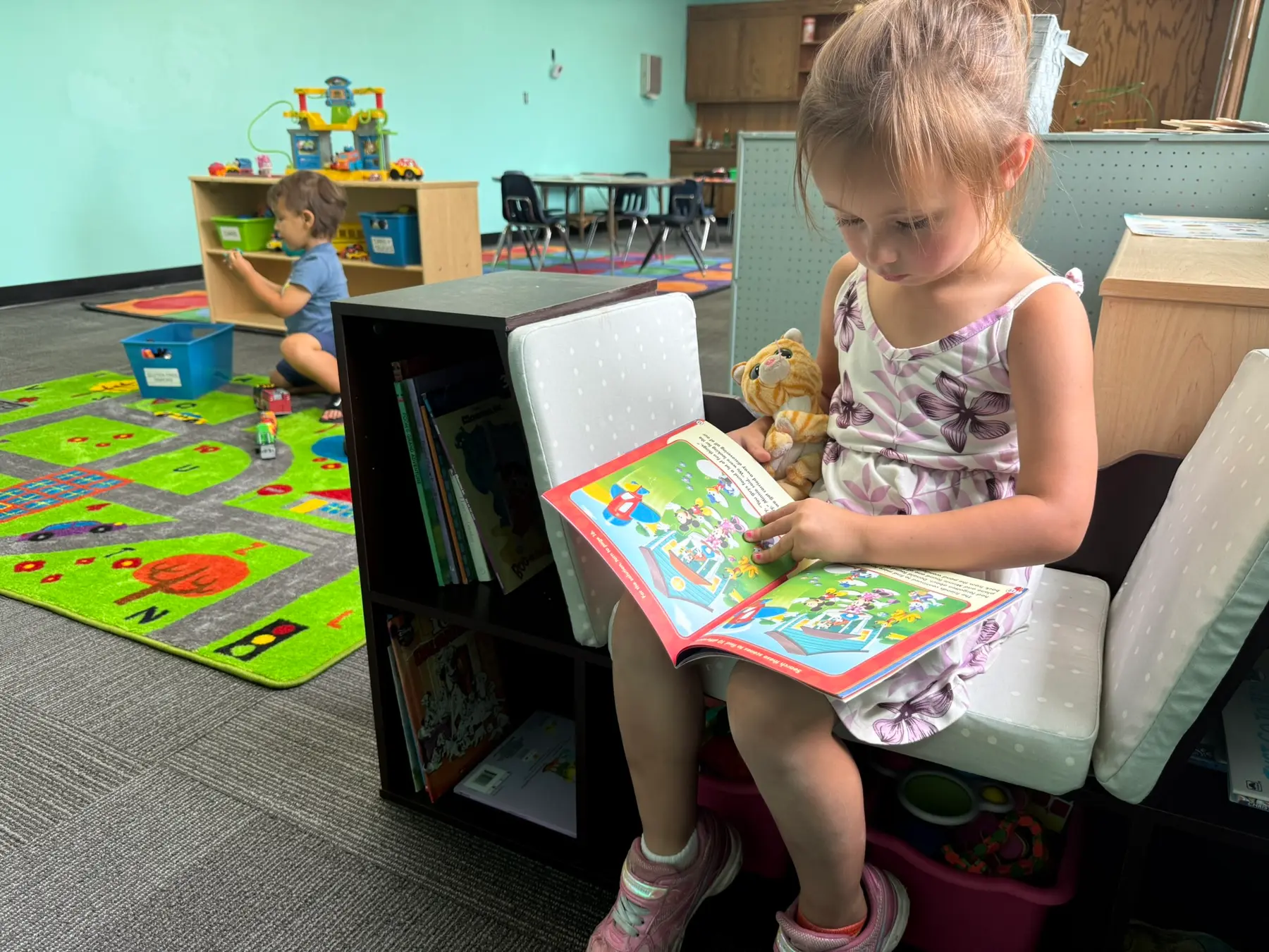 A girl in a floral dress sits on a white chair reading a book while holding a stuffed tiger, and a boy sits on the floor near a shelf with toys.