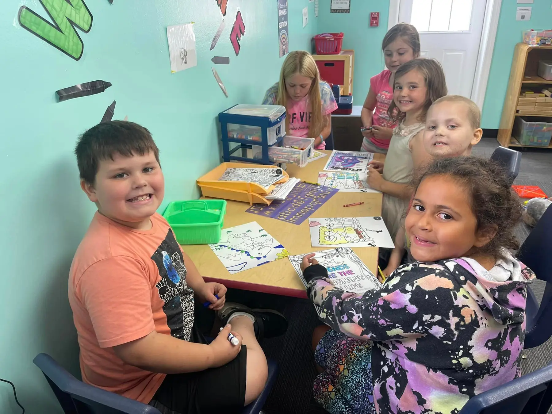 Six children sit and stand around a table coloring pages with crayons in a classroom with teal walls.