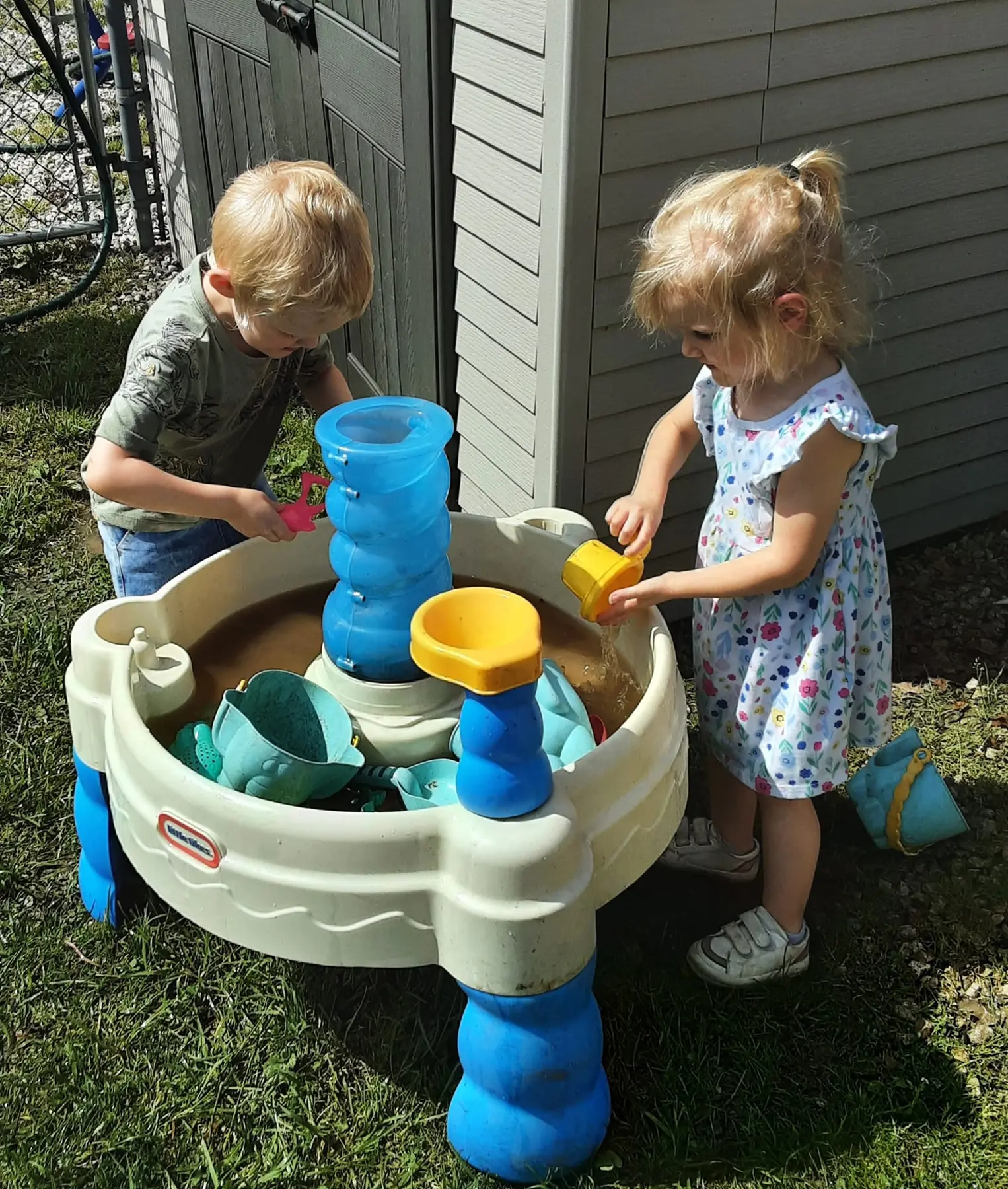 Two young children stand at a plastic water play table, one pouring water from a yellow cup while the other holds a pink toy.