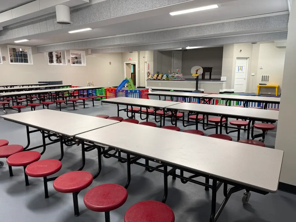 An empty multi-purpose room with long folding tables attached to red stools, colorful storage bins, and a small stage area in the background.