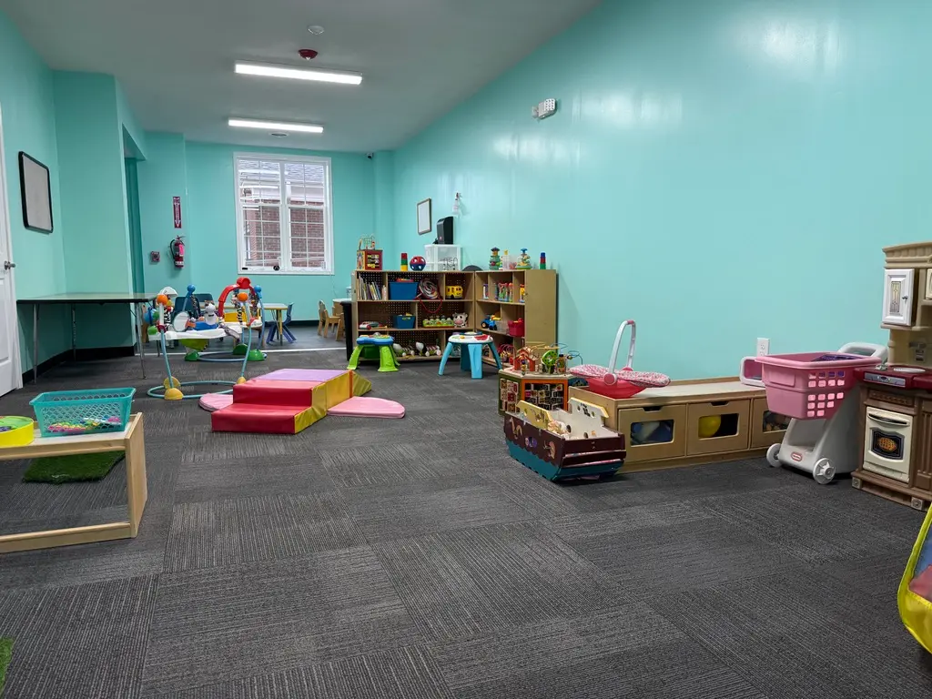 A brightly lit childcare room with teal walls, gray carpet, and various play areas including a soft foam climber, toy kitchen, shelves of toys, and small tables.