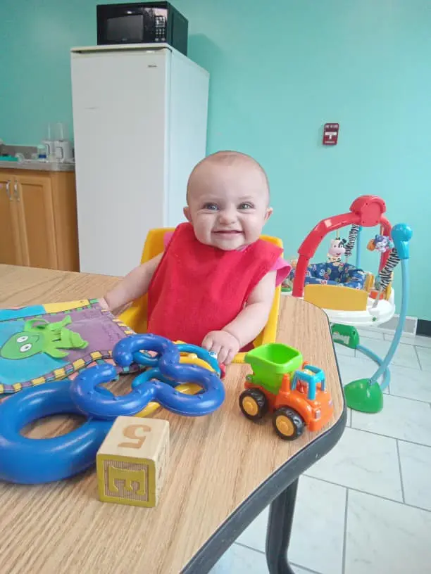 A baby wearing a red bib sits in a yellow seat at a table with a book, blue toy links, a block, and a toy tractor.