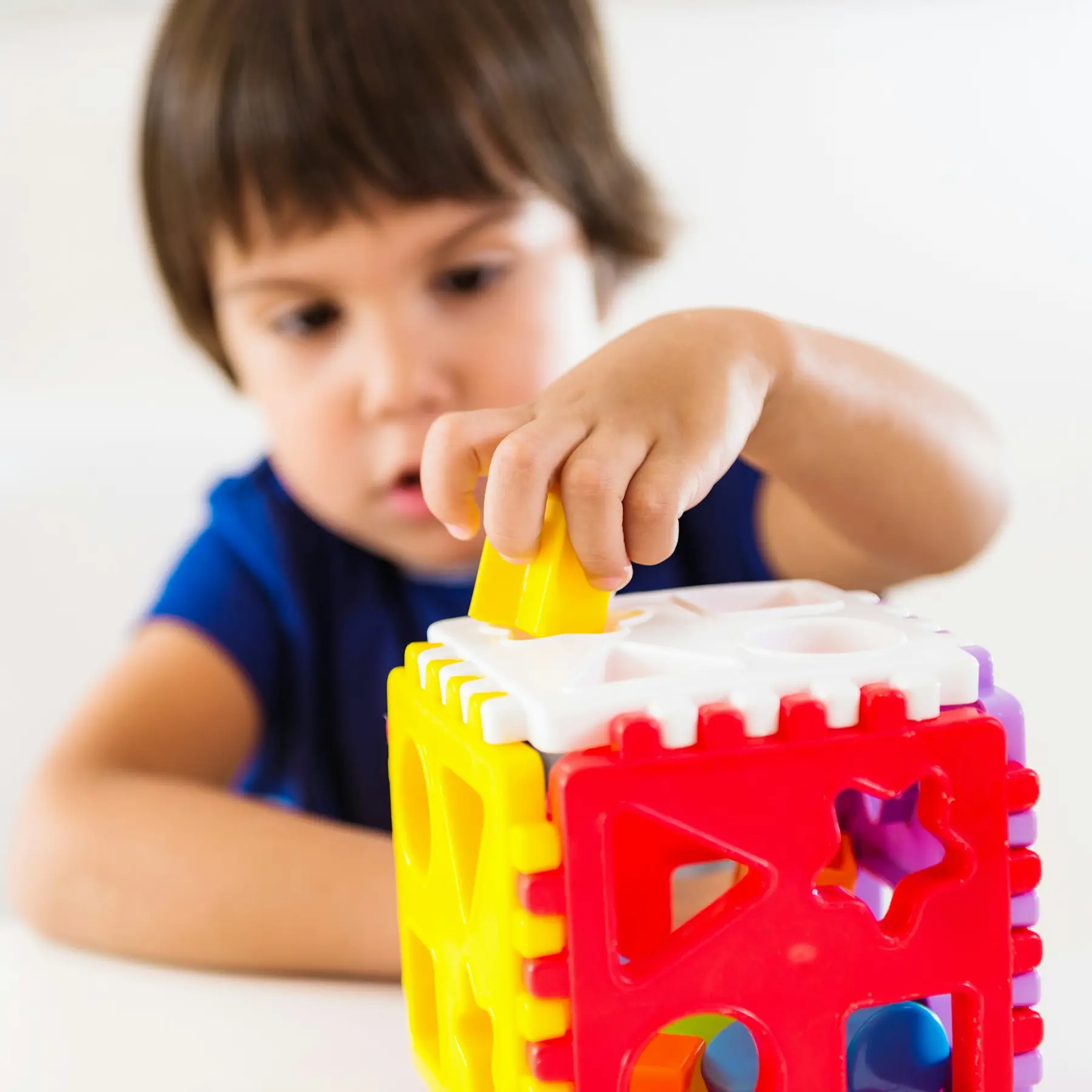 A young child in a blue shirt places a yellow shape into a colorful plastic shape sorter toy on a white surface.