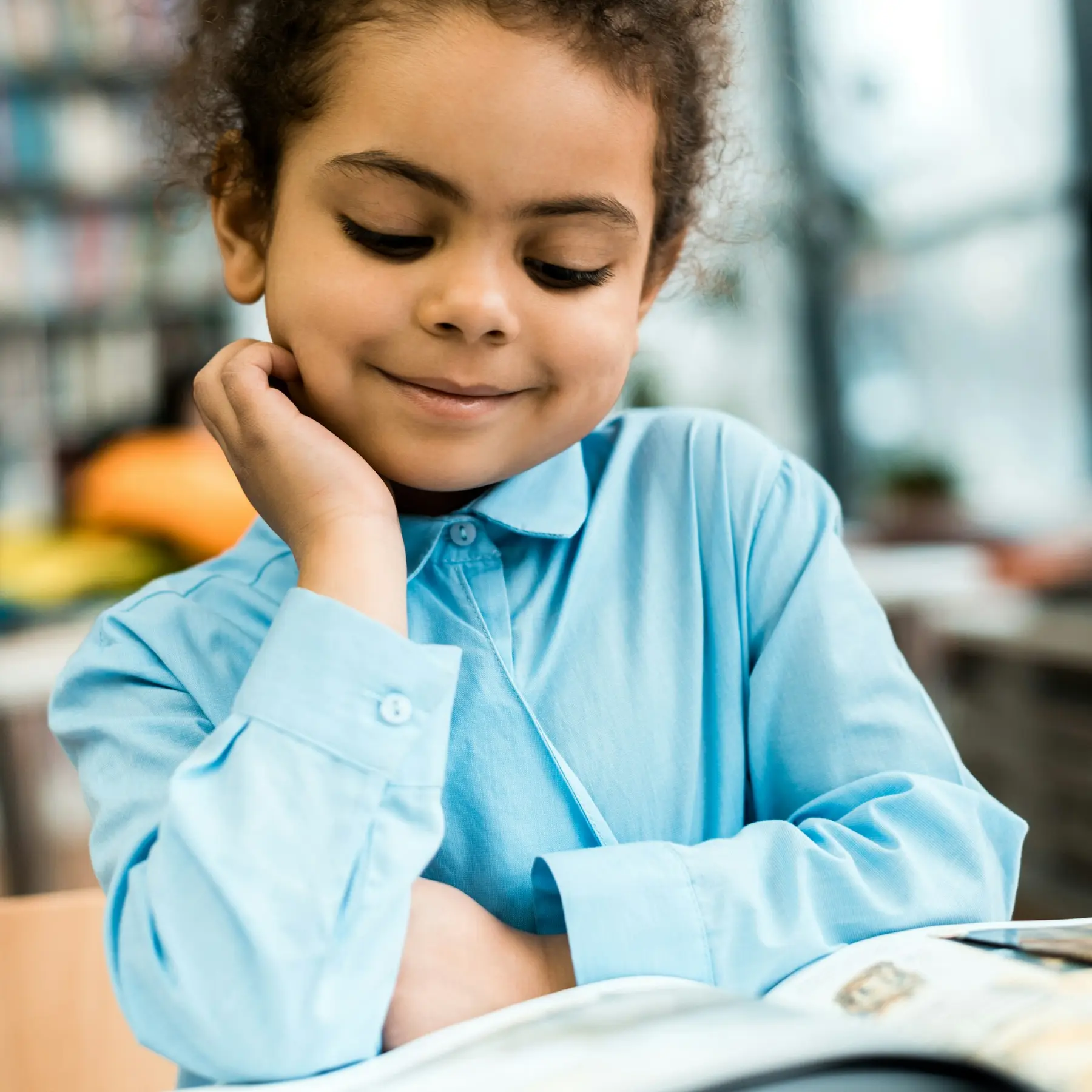 A young girl in a light blue button-up shirt rests her chin on her hand while smiling and looking down at an open book.
