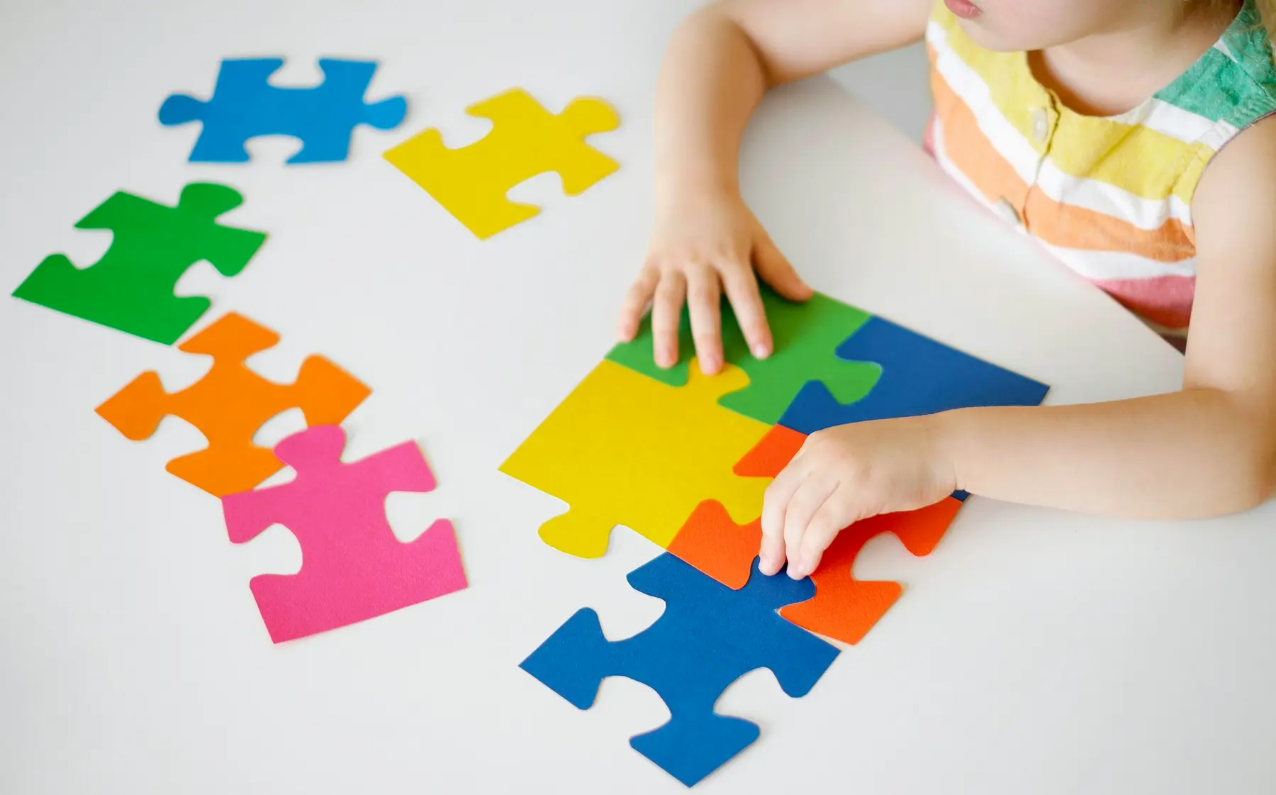A child in a striped top places colorful jigsaw puzzle pieces on a white table.