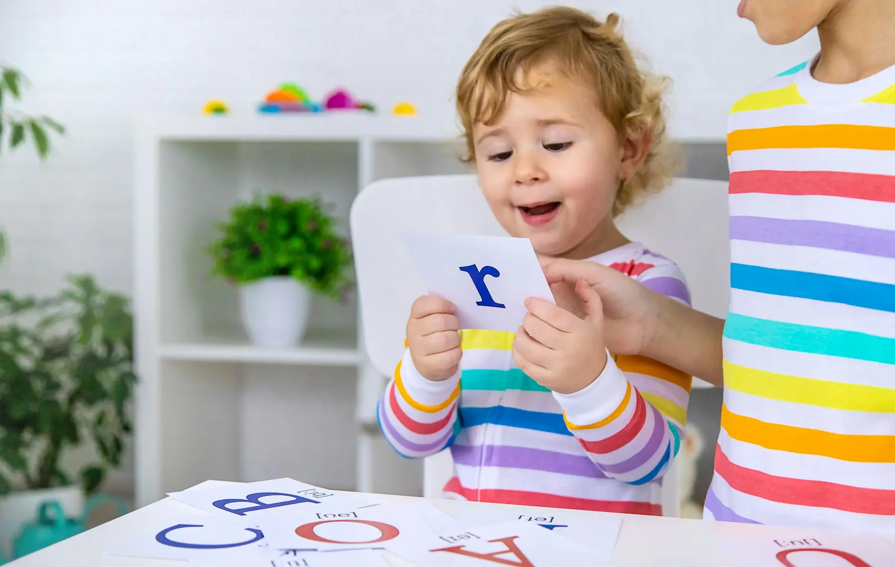 A toddler in a striped shirt holds up a card with a blue letter r while another person points at it, with alphabet cards on a table in front of them.