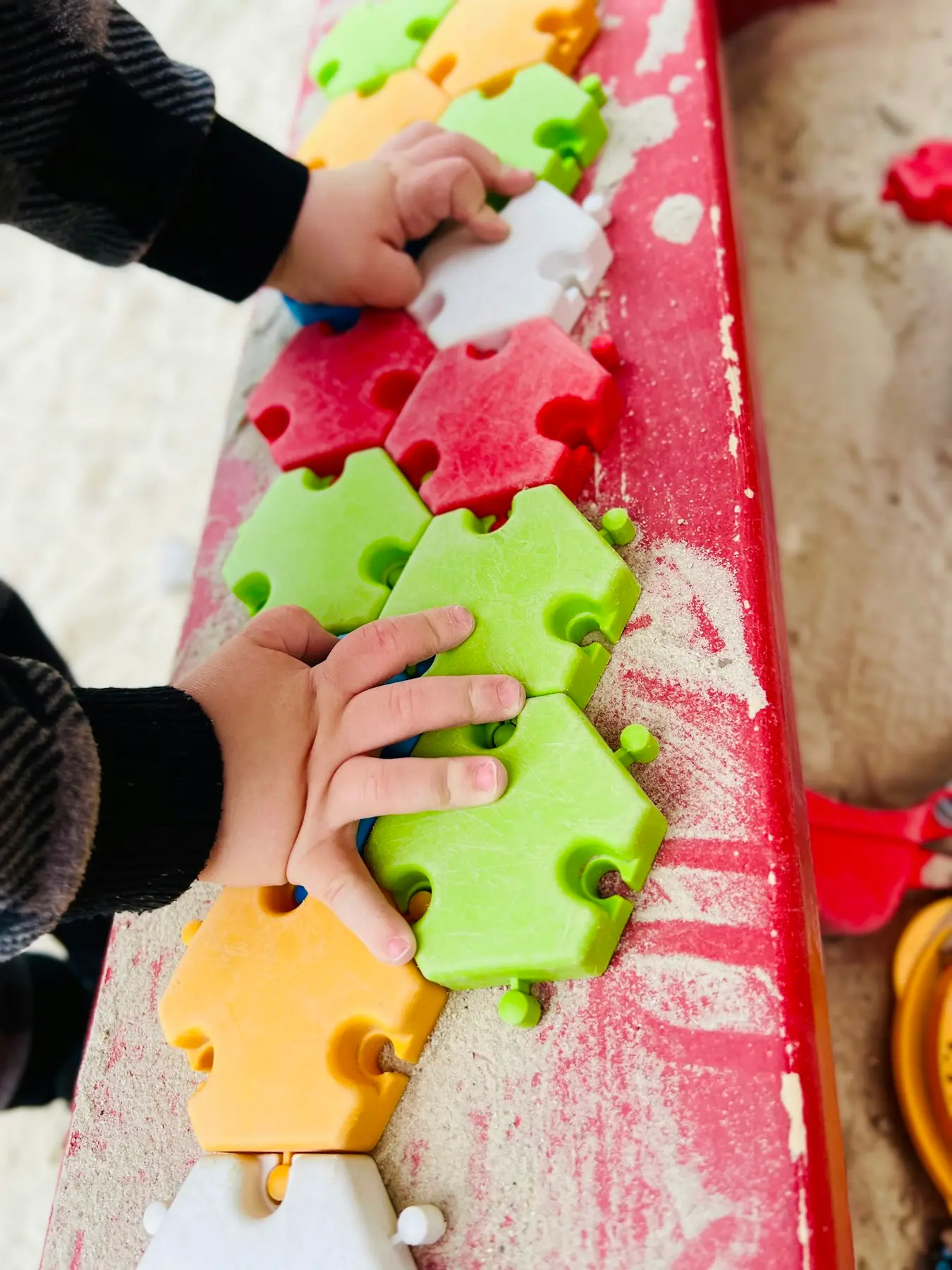 A child wearing a black long-sleeved shirt uses both hands to connect colorful interlocking foam puzzle tiles on a red surface covered with scattered sand.