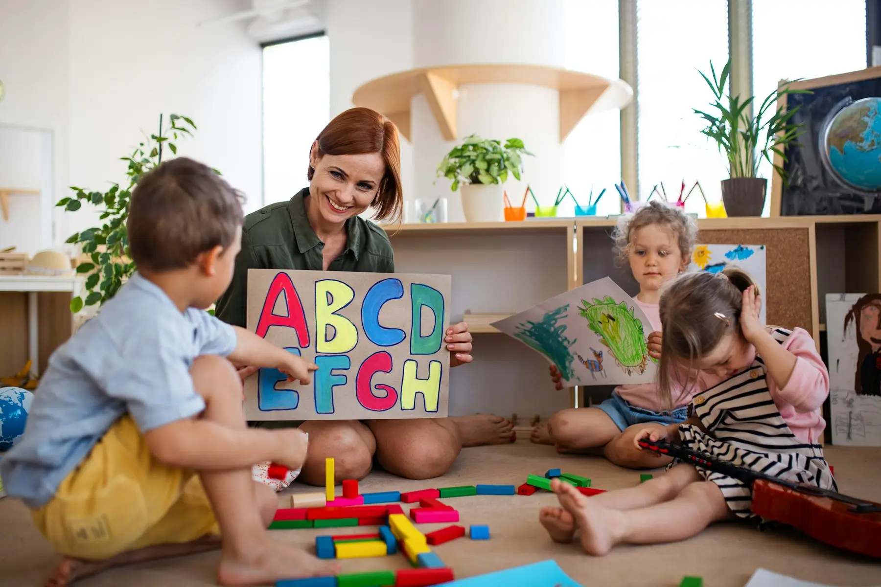 A woman holds an alphabet sign while three children sit on the floor with blocks, a drawing, and a small guitar.