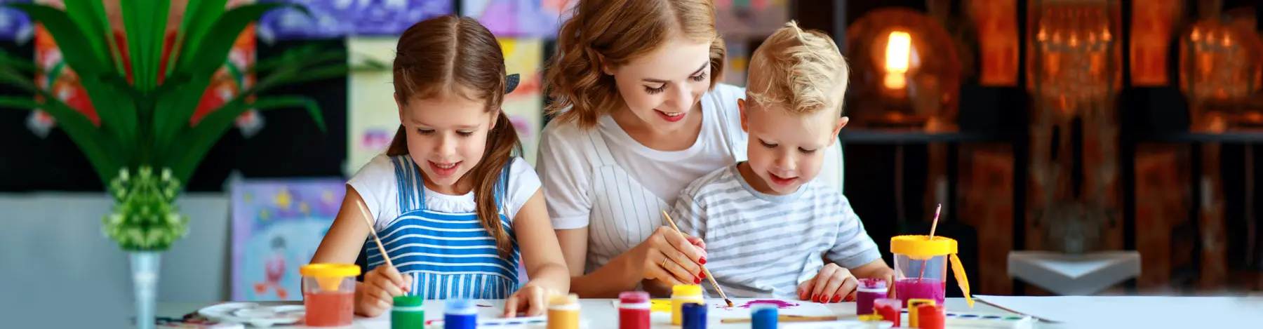 A woman sits between two children at a table, helping them paint with brushes and colorful cups of paint.