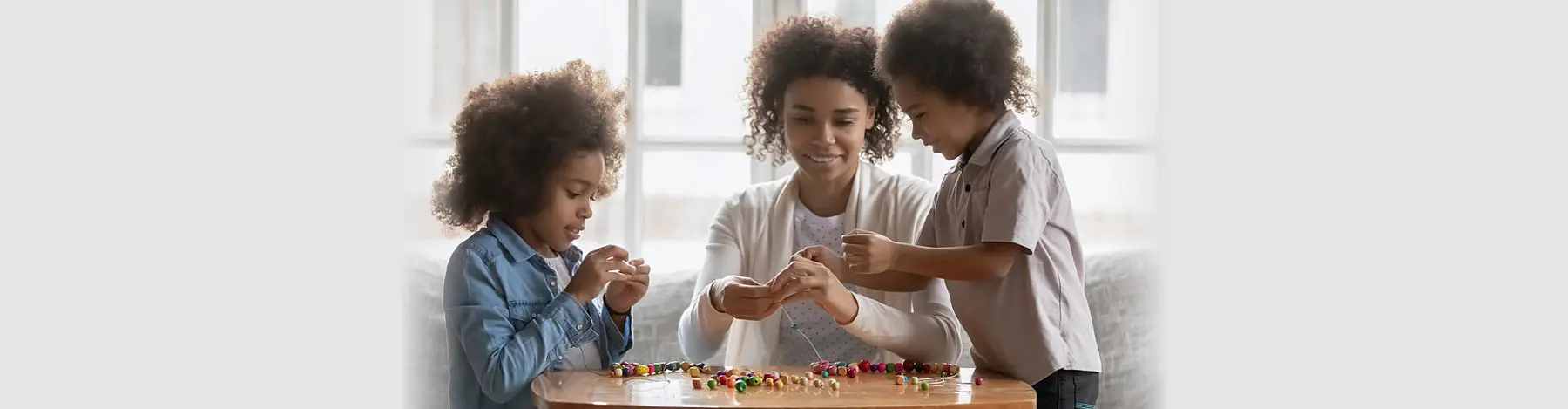 A woman and two children sit at a wooden table and handle small colorful craft pieces.
