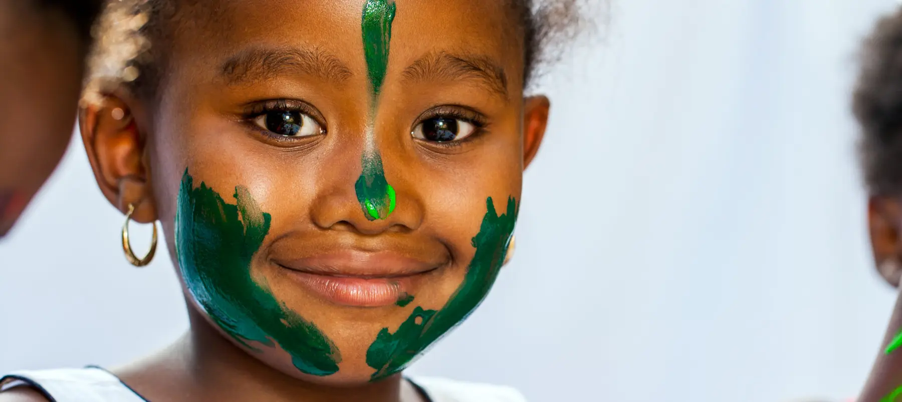 A young girl with green face paint smiles at the camera while two other children are partially visible at the edges of the frame.
