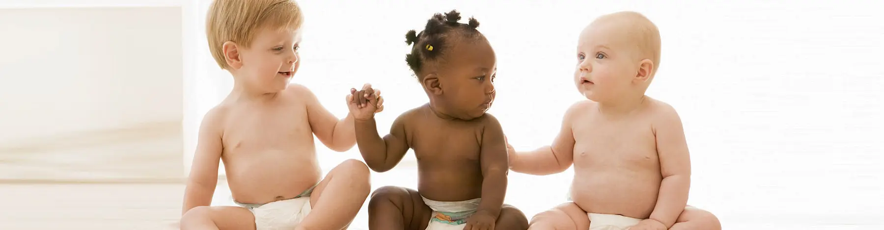 Three babies wearing diapers sit together on a light floor, with the baby on the left holding hands with the baby in the middle while the baby on the right reaches toward them.