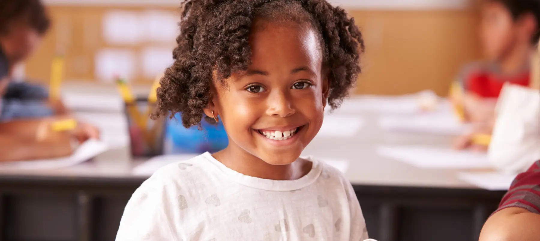 A smiling girl with curly hair in a white patterned shirt sits at a table while other children write on papers in the background.