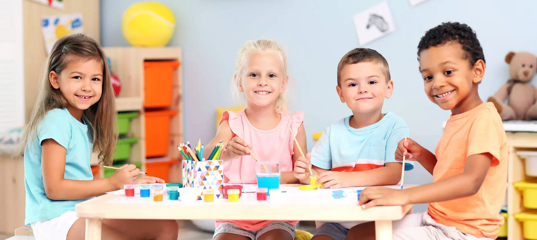 Four children sit at a table painting with brushes and small cups of paint in a classroom.