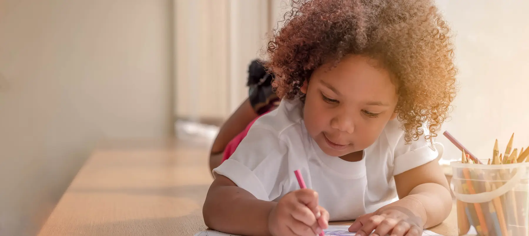 A young child with curly hair lies on a wooden surface, drawing on paper with a pink pencil while a container of colored pencils sits nearby.