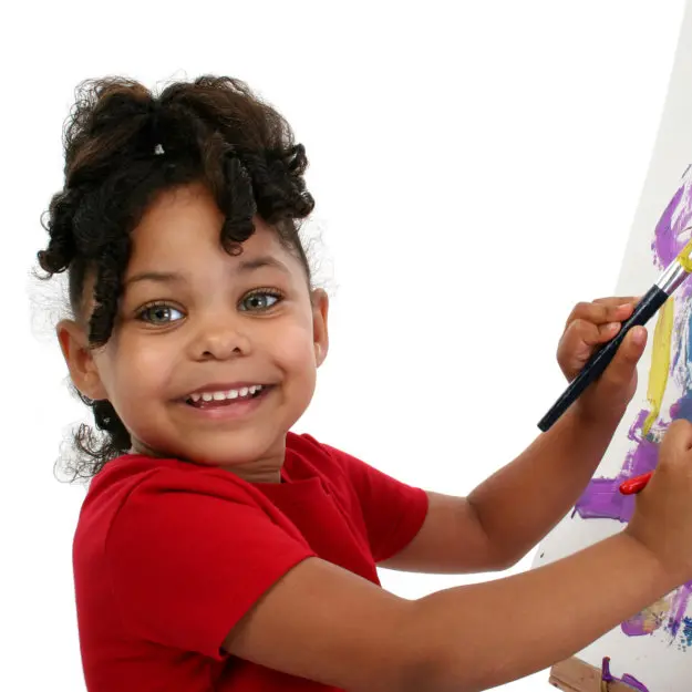 A smiling girl in a red shirt holds a paintbrush and paints on a white easel.