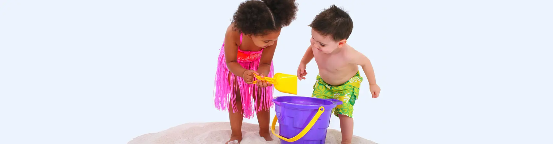 Two young children play with a yellow shovel and a purple bucket on a small pile of sand against a plain background.