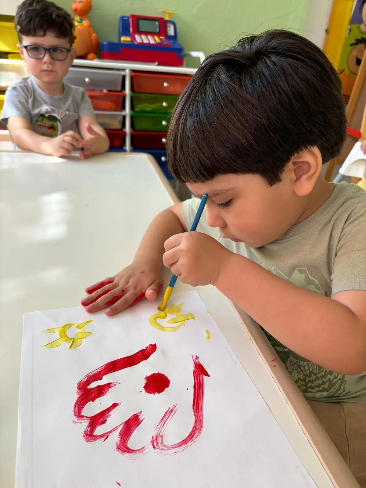 A boy in a green shirt paints red and yellow strokes on paper with a brush while another boy in glasses sits at the table behind him.