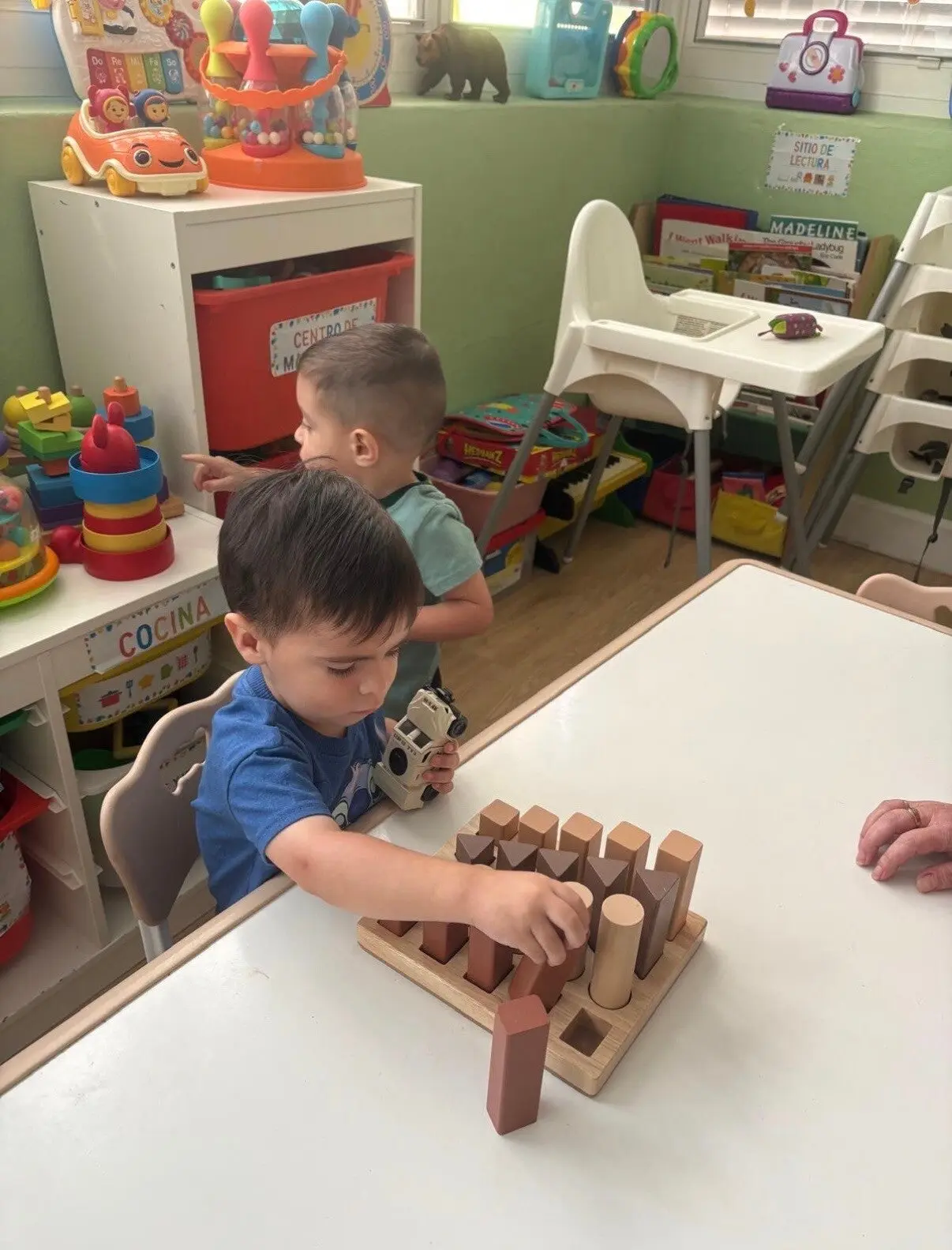 A boy in a blue shirt sits at a table placing wooden blocks into a shape sorter while another boy stands behind him pointing, in a classroom with toys and shelves.